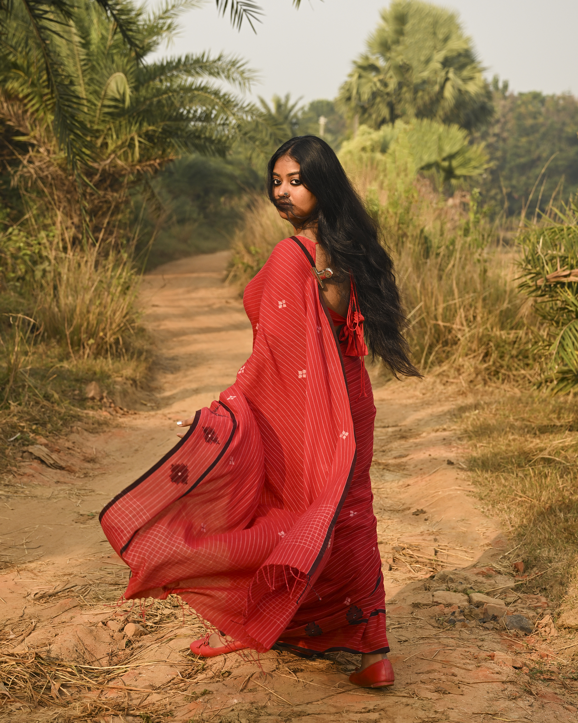 Model wearing the Vermilion Linear Jamdani Saree, a handwoven vermilion red with black-edged borders Jamdani saree with fine linear rhythm with scattered woven motifs and graphic pallu with sharp contrast-led edging.